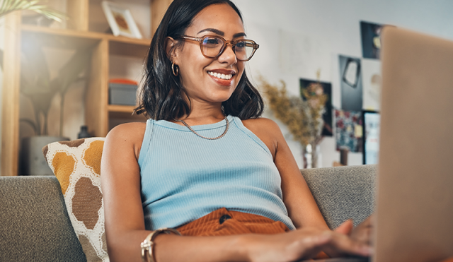 Photo of young woman on laptop sitting on a couch in a blue shirt and orange pants 