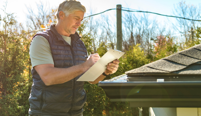A man stands on a ladder and holds a clipboard near a roof