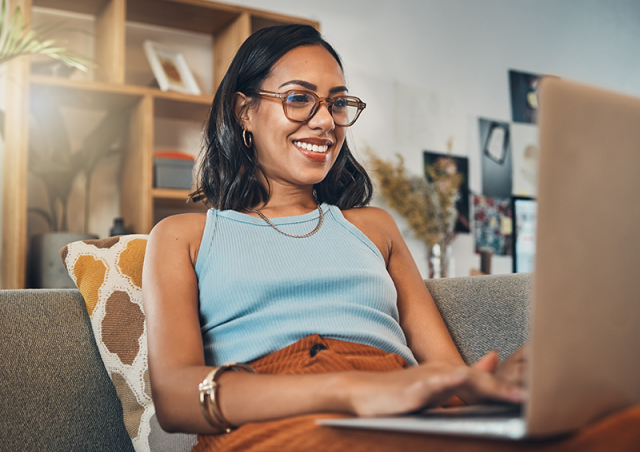 Photo of young woman on laptop sitting on a couch in a blue shirt and orange pants 