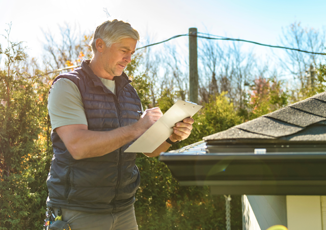A man stands on a ladder and holds a clipboard near a roof